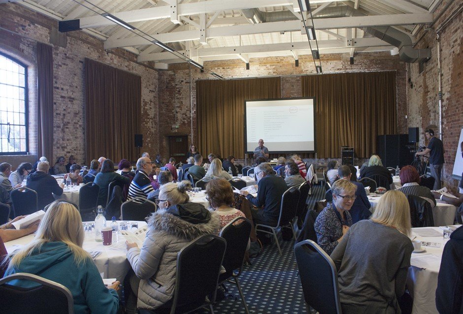 Photograph of a hall with lots of tables and people seated around them, looking at a presenter at the front of the room who is stood in front of a whiteboard