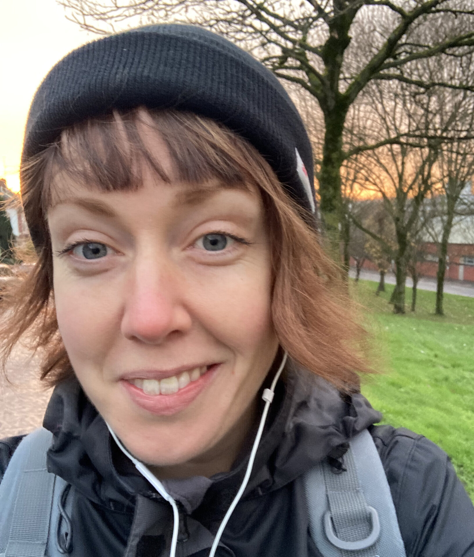 Selfie of Jennifer wearing a black hat, a black coat, and white earphones in front of a tree where the sunset is visible through the branches