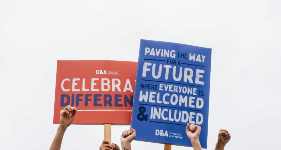 Arms raised to the sky, either with their hands in fists or holding up protest boards. One protest board is red, and reads “Diversity and Ability- celebrate difference.” The other is blue, and reads “Paving the way for a future where everyone is welcomed and included”.