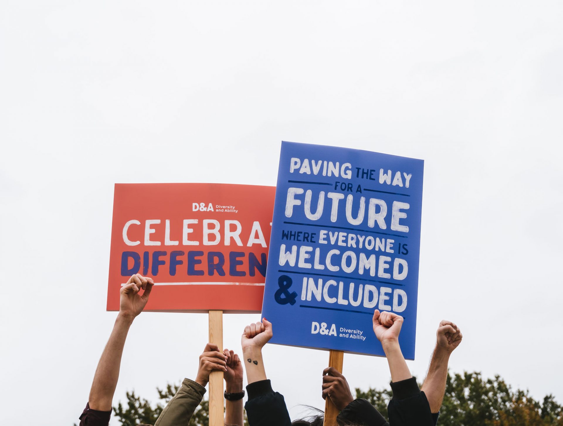 Arms raised to the sky, either with their hands in fists or holding up protest boards. One protest board is red, and reads “Diversity and Ability- celebrate difference.” The other is blue, and reads “Paving the way for a future where everyone is welcomed and included”.