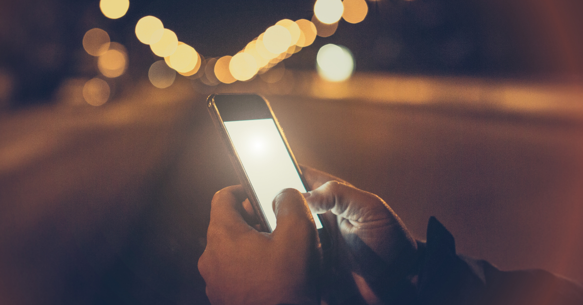 A picture of a pair of hands holding a brightly-lit smartphone. The backdrop is a street at night lined with blurry streetlamps.