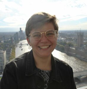 A headshot of Ave smiling at the camera with the Thames in the background. Ave has short light brown hair and wears gold rimmed glasses.
