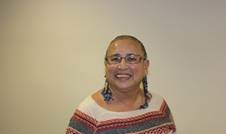 A photo of June, who wears glasses and is wearing a white, red and grey patterned jumper against a plain beige background