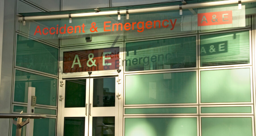 The front of a hospital. The text Accident & Emergency is visible in red, above light green doors and the wording A & E in big white lettering against red.